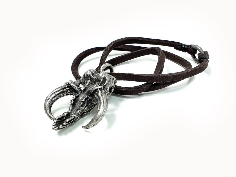 May include: A silver pendant shaped like a claw with a brown leather cord. The pendant has a detailed design and is a unique piece of jewelry.