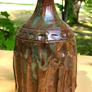Peut inclure: Un vase en céramique fait à la main avec une surface brillante et texturée. Le vase présente une palette de couleurs marron et sarcelle avec une bande décorative d'indentations circulaires. Le vase a un col étroit et un corps arrondi.