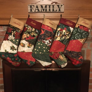Puede incluir: Cinco calcetines navideños personalizados cuelgan sobre una chimenea. Cada calcetín presenta un diseño de patchwork con telas rojas, verdes y crema, adornadas con motivos florales y navideños. Un letrero de madera arriba dice "FAMILY".