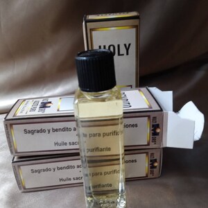 May include: A clear glass bottle with a black cap containing a light yellow liquid, positioned in front of three rectangular cardboard boxes. The boxes feature white labels with black text including the words HOLY, BENDITO, and ACEITE PURO.