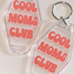 以下が含まれることがあります： コーラル色の文字で「COOL MOMS CLUB」と書かれた2つの透明なアクリルキーホルダー。キーホルダーはレトロなモーテルのキーデザインで、シルバーのキーリングが付いています。背景は明るくニュートラルな色です。