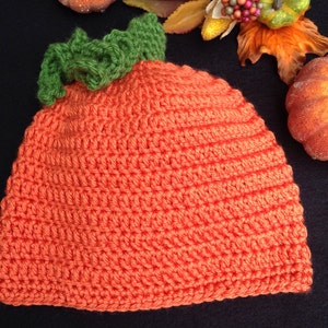 May include: Orange crocheted pumpkin hat with a green crocheted stem. The hat is on a black surface.