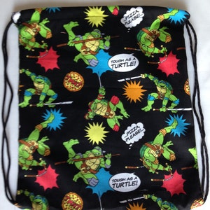 K&ouml;nnte beinhalten: Schwarze Kordelzugtasche mit Teenage Mutant Ninja Turtles-Motiv. Die Tasche zeigt ein sich wiederholendes Muster der Turtles in Gr&uuml;n, Schwarz und Rot, sowie Pizzast&uuml;cke und den Text "Tough as a Turtle!" und "...Pizza, Please!"