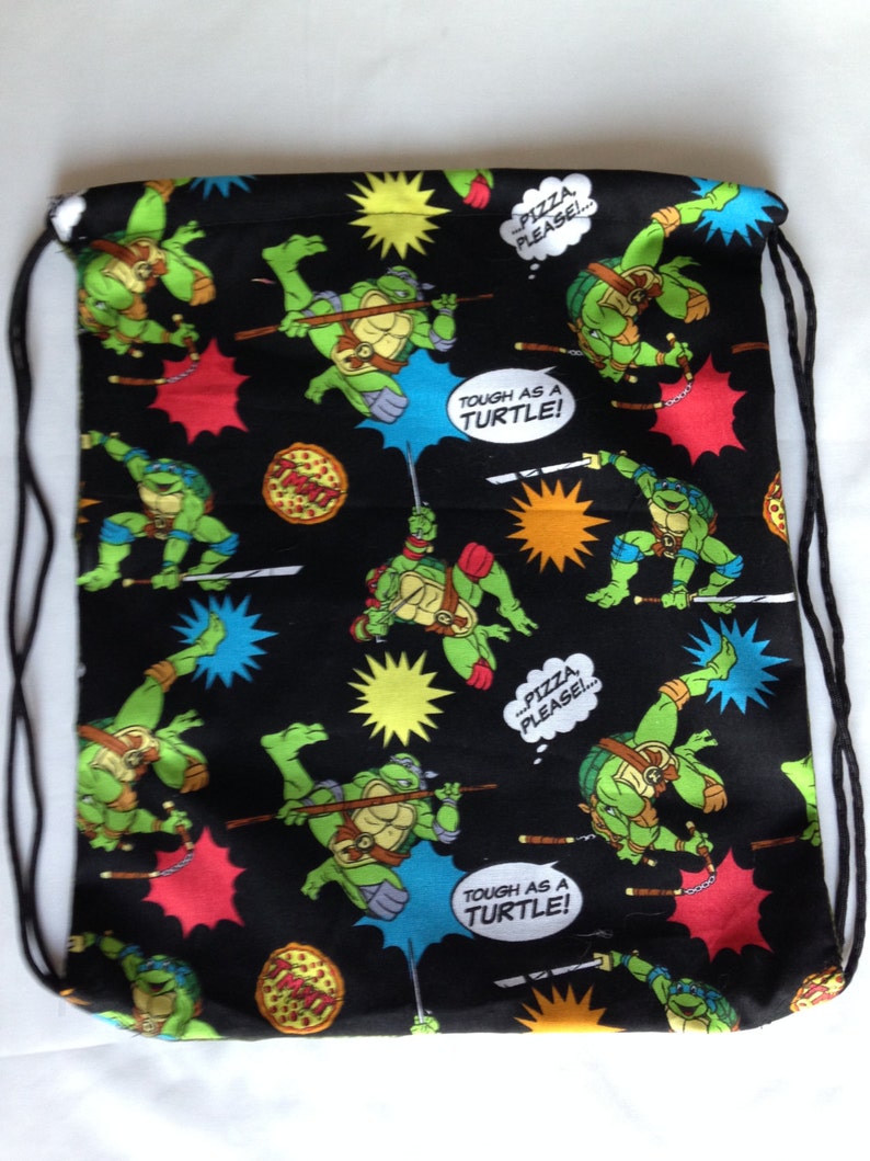 K&ouml;nnte beinhalten: Schwarze Kordelzugtasche mit Teenage Mutant Ninja Turtles-Motiv. Die Tasche zeigt ein sich wiederholendes Muster der Turtles in Gr&uuml;n, Schwarz und Rot, sowie Pizzast&uuml;cke und den Text "Tough as a Turtle!" und "...Pizza, Please!"