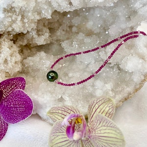 May include: A delicate pink gemstone necklace with a single dark green pearl pendant. The necklace is displayed on a white crystal geode with two pink and white orchids.