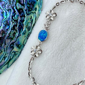 May include: A silver chain bracelet with three white flower charms and a blue opal stone. The bracelet is on a white fluffy surface.
