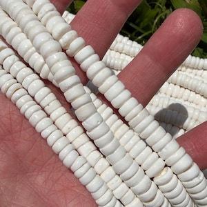May include: Close-up of several strands of white, disc-shaped beads. The beads are uniform in size and shape, with a slightly rounded edge. The strands are held in a hand, with green grass visible in the background. The beads appear to be made of a natural material.