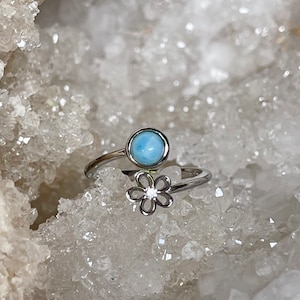 May include: A silver ring with a blue gemstone and a flower design. The ring is set on a white crystal background.