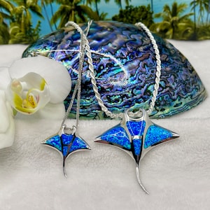 May include: Two silver necklaces with stingray pendants. The pendants feature a vibrant blue opal inlay and are suspended from a silver rope chain. The stingray pendants are displayed on a white surface with a decorative shell and a white orchid flower in the background.