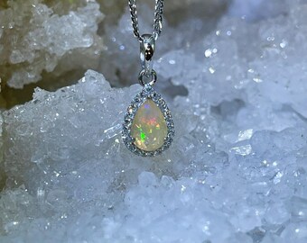 May include: A pear-shaped opal pendant with a silver chain and a halo of small clear stones. The pendant is set against a background of white crystals.