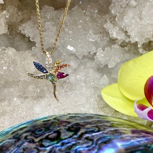 May include: A gold necklace with a colorful hummingbird pendant. The pendant is adorned with various gemstones, including red, blue, green, and yellow.