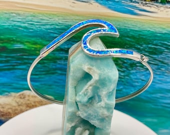 May include: A silver-toned wave-shaped bracelet with blue opal inlays. The bracelet is displayed on a light blue stone, with a tropical beach scene in the background. A white plate and purple flower petals are in the foreground.