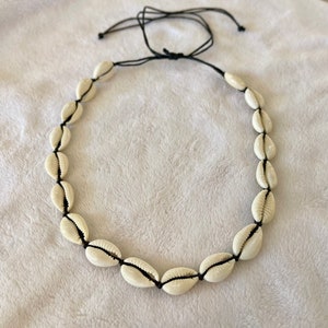 May include: A necklace featuring a series of cream-colored seashells strung together with black cord. The shells are evenly spaced and the cord is tied at the back for an adjustable fit. The necklace is a beach-themed accessory.