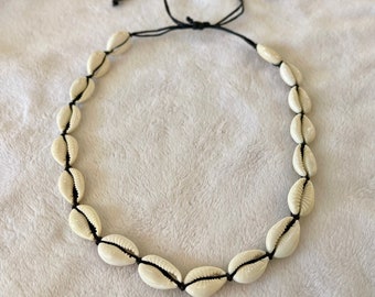 May include: A necklace composed of cream-coloured seashells, threaded onto a black cord. The shells are uniformly spaced, and the cord is tied at the back for adjustment. This necklace is a beach-themed accessory, perfect for summer.