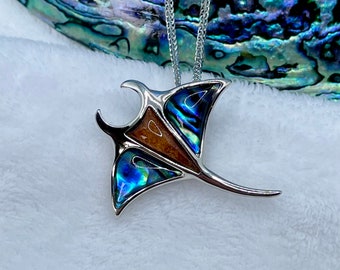 May include: A silver-toned pendant necklace featuring a stylized manta ray design. The pendant incorporates iridescent abalone shell and a brown inlay, suspended from a delicate chain. The background features a blurred iridescent surface.