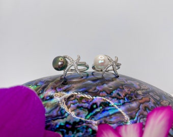 May include: Two silver rings with a starfish design and a pearl. The ring on the left has a black pearl and the ring on the right has a white pearl. The rings are sitting on a shell with a purple flower in the foreground.