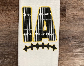 May include: White waffle-weave kitchen towel with a football field design. The design features the letters "IA" in gold with a black and white football field pattern. The bottom of the design has a gold football stitch pattern.
