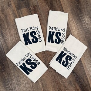 May include: Four white kitchen towels with black embroidered text. Each towel features a different Kansas city name and zip code. The cities are Fort Riley, Milford, Junction City, and Manhattan.