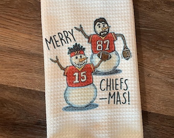 May include: White kitchen towel with a red, white, and blue snowman design. The snowman is wearing a red and white football jersey with the number 15. The text "Merry Chiefs-Mas!" is printed on the towel.