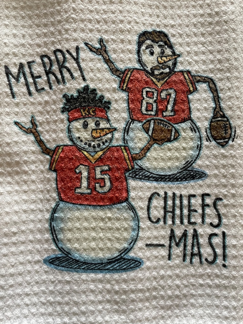 May include: A white kitchen towel with a red and white snowman wearing a red jersey with the number 15 and the letters "KC".  The snowman is holding a football.  Behind the snowman is another snowman wearing a red jersey with the number 82.  The text "Merry Chiefs-Mas!" is written on the towel.