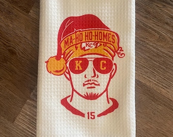 May include: White kitchen towel with a red and yellow illustration of a person wearing a Santa hat and sunglasses. The text "MA-HO HO-HOMES" is on the hat and "KC" is on the sunglasses. The number "15" is below the person's face.