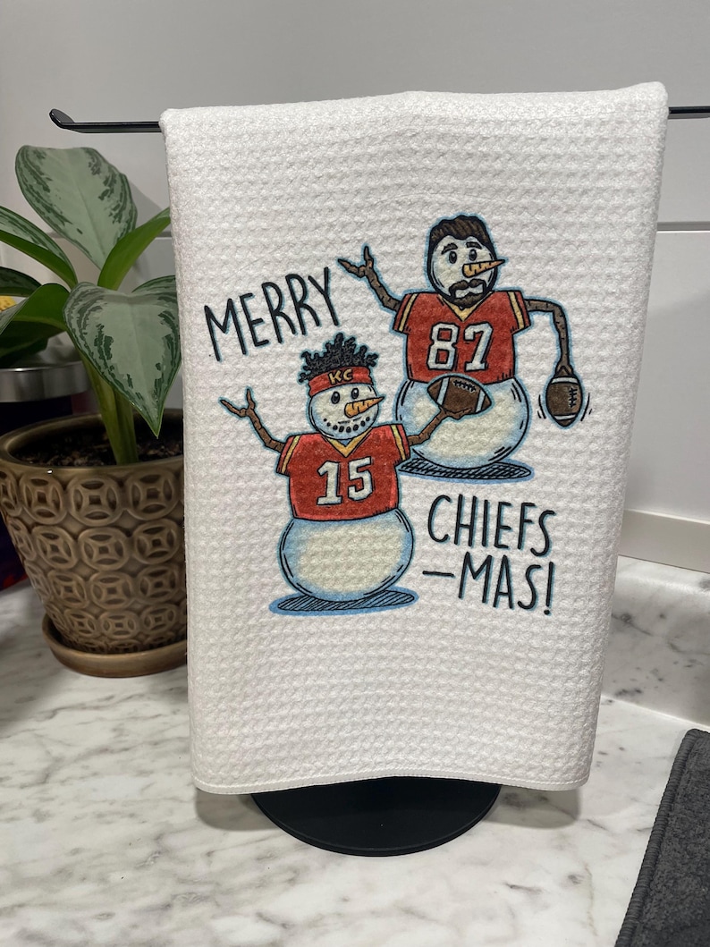 May include: White kitchen towel with a red and white snowman design. The snowman is wearing a red jersey with the number 15 and the letters "KC". The text "Merry Chiefs-Mas!" is printed on the towel.