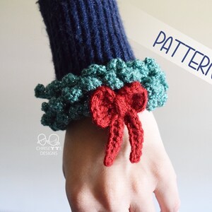 May include: A close-up of a crocheted green and teal wreath with a red bow. The wreath is worn on a person's wrist as a bracelet. The word "PATTERN" is written in blue text on a white background.