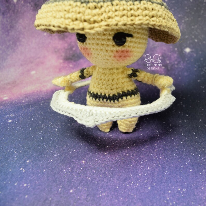 May include: A crocheted amigurumi toy shaped like a planet with a ring system. The toy is beige with black accents and has a wide-brimmed hat. The ring system is white and is made of crocheted yarn. The toy is posed against a purple and white galaxy background.