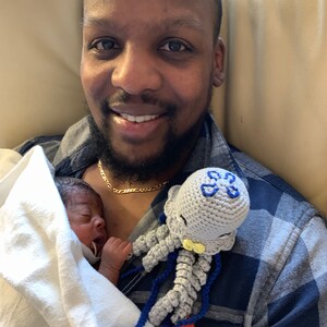 May include: A man is holding a newborn baby wrapped in a white blanket. The baby is wearing a white onesie. The man is wearing a blue and black plaid shirt and a gold chain. A gray crocheted jellyfish toy with blue tentacles is on the man's shoulder.
