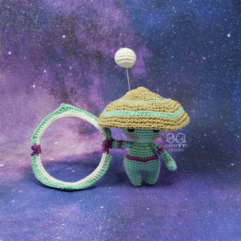 May include: A crocheted alien doll wearing a brown and green hat with a white ball on top. The doll is holding a green and white hoop. The doll is standing on a purple and blue background.