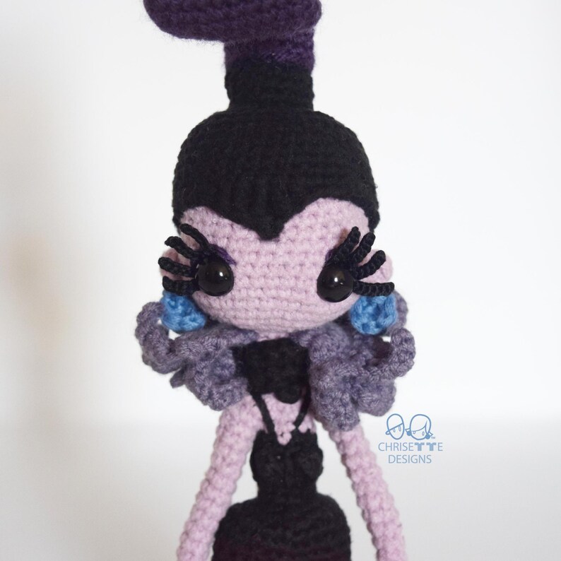 May include: A handmade crochet doll with a purple and black color scheme. The doll has large black eyes, long eyelashes, and blue accents. The doll's hair is black, and the skin is lavender. The doll is wearing a black dress.