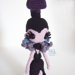 May include: A handmade crochet doll in a gothic style. The doll features a purple headpiece, black hair, and a lavender face with large black eyelashes. It wears a black dress with a lavender collar and blue accents. The doll is a unique, handcrafted item.