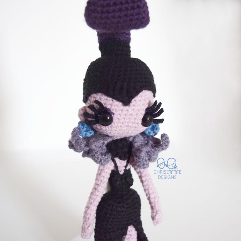 May include: A handmade crochet doll in a gothic style. The doll features a purple headpiece, black hair, and a lavender face with large black eyelashes. It wears a black dress with a lavender collar and blue accents. The doll is a unique, handcrafted item.