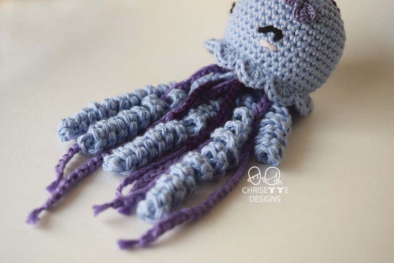 May include: A crocheted jellyfish toy with a light blue body and purple tentacles. The jellyfish has a cute face with embroidered eyes and a pink blush.