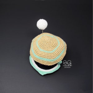 May include: A crocheted hat with a light brown base and a light green band. The hat has a white pom-pom on top. The hat is sitting on a black surface.  Chrissette Designs.