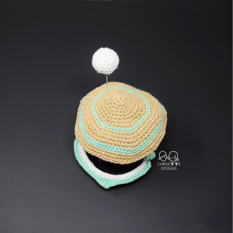 May include: A crocheted hat with a light brown base and a light green band. The hat has a white pom-pom on top. The hat is sitting on a black surface.  Chrissette Designs.