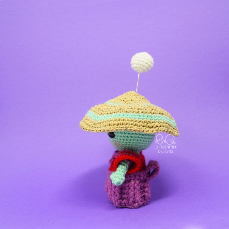 May include: A crocheted amigurumi doll wearing a green and brown hat with a white pom-pom on top. The doll is wearing a red and purple outfit. The doll is standing on a purple background.  Chrisette Designs.