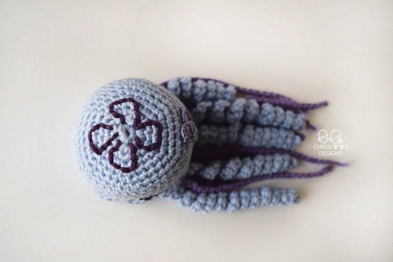 May include: A crocheted octopus toy with a light blue body and purple tentacles. The octopus has a purple four-leaf clover design on its head.