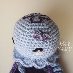 May include: A crocheted octopus amigurumi toy with a light blue body, purple tentacles, and a purple bow on its head. The octopus has black eyes and a black mustache.