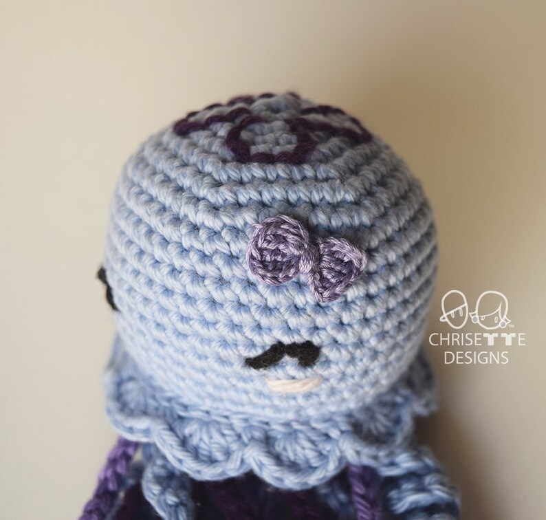 May include: A crocheted octopus amigurumi toy with a light blue body, purple tentacles, and a purple bow on its head. The octopus has black eyes and a black mustache.