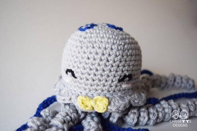 May include: A crocheted gray octopus with blue tentacles and a yellow bow tie. The octopus has a cute face with black eyes and a pink blush.