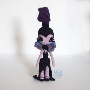 May include: Crocheted doll in a black dress with a high slit, purple hair, and blue accents. The doll has a black bodice and a long, flowing skirt. The doll's face is light purple with black eyelashes and blue earrings. The doll is handmade.