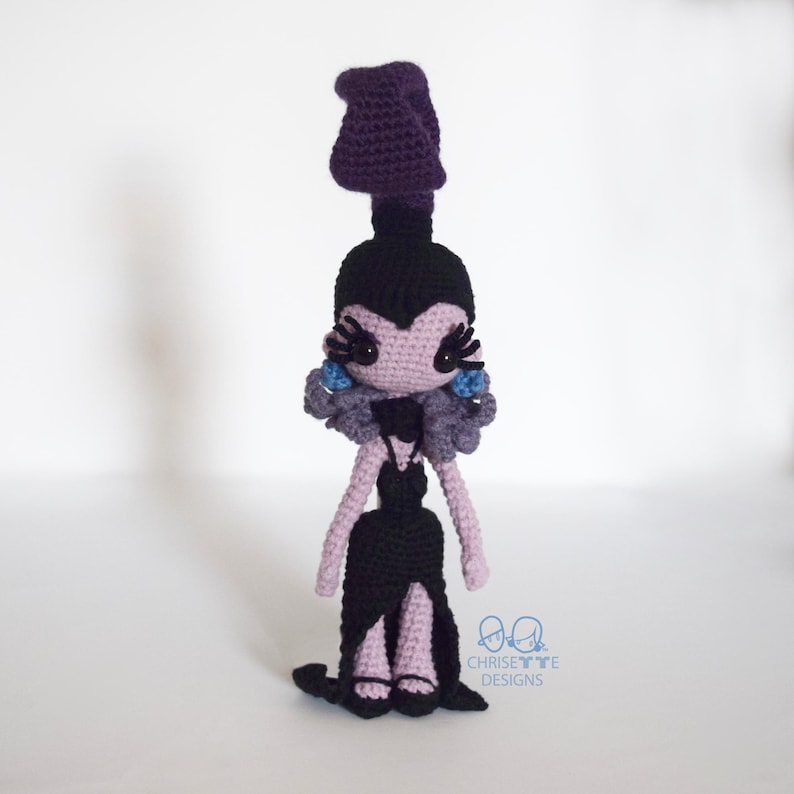 May include: Crocheted doll in a black dress with a high slit, purple hair, and blue accents. The doll has a black bodice and a long, flowing skirt. The doll's face is light purple with black eyelashes and blue earrings. The doll is handmade.