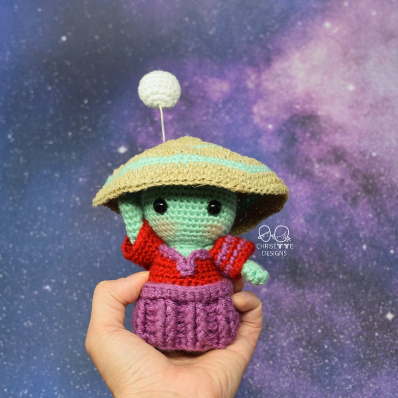 May include: A crocheted alien doll wearing a red and purple dress and a brown and green hat with a white ball on top. The doll is being held in a hand against a purple and blue starry background.