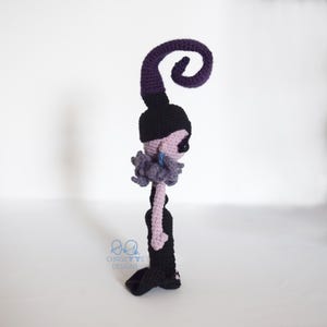 May include: Crocheted doll in a side profile. The doll has a black hat with a purple swirl, a black dress, and a light purple face and arms. The doll has a gray ruffled collar and a blue earring. The doll is handmade.