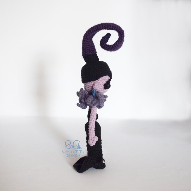 May include: Crocheted doll in a side profile. The doll has a black hat with a purple swirl, a black dress, and a light purple face and arms. The doll has a gray ruffled collar and a blue earring. The doll is handmade.