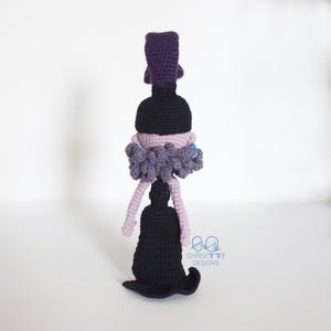May include: A handmade crochet doll in a witch design. The doll features a black hat, a black dress, and a purple and gray ruffled collar. The doll has light purple legs and a black base. The doll is standing upright.