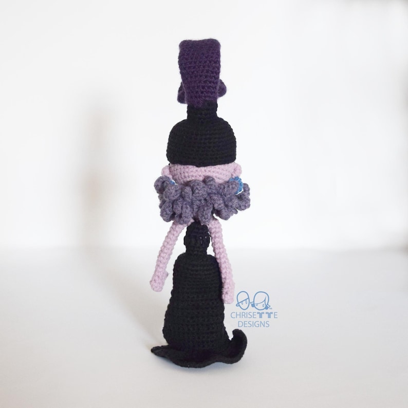 May include: A handmade crochet doll in a witch design. The doll features a black hat, a black dress, and a purple and gray ruffled collar. The doll has light purple legs and a black base. The doll is standing upright.