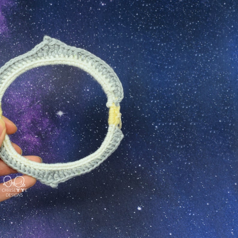 May include: A crocheted ring with a white and gray color scheme. The ring is held in a hand against a dark blue background with white stars.