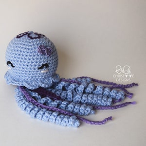 May include: A crocheted blue jellyfish with purple tentacles and a purple bow on its head. The jellyfish has embroidered eyes and a small flower on its head.